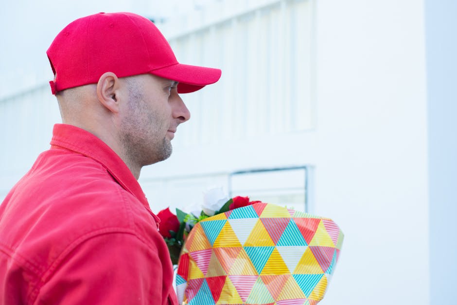 A man dressed in a red cap and jacket holding a floral gift wrapped in colorful geometric patterned paper with a ribbon, standing outdoors against a light-colored building and a cloudy sky background.