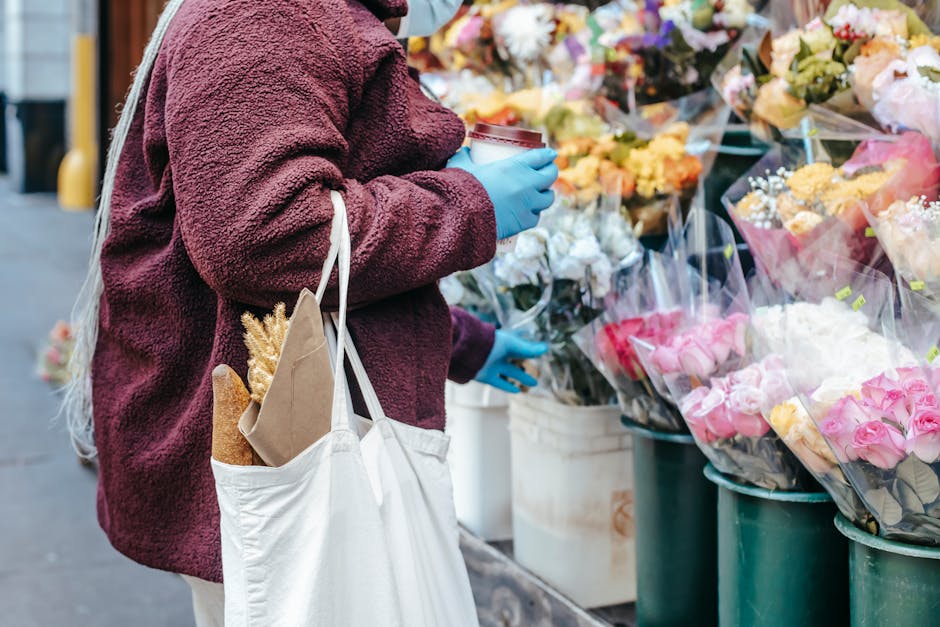 A person wearing a burgundy fleece jacket and blue gloves is shopping for fresh flowers at an outdoor flower stall near the Harvey Centre. They are holding a white cloth tote bag containing a baguette