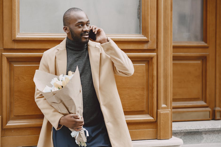 A man with a dark complexion and a neatly trimmed beard, dressed in a light beige coat over a dark grey turtleneck, is standing outdoors in front of a wooden panelled building. He is holding a bouquet