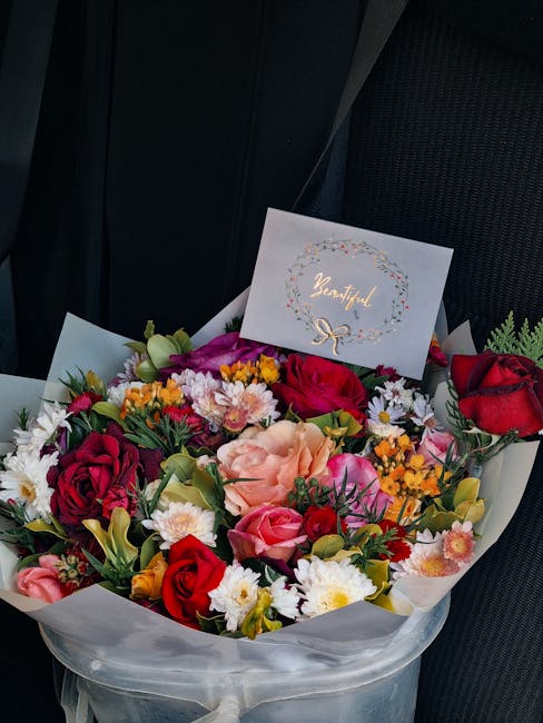 A vibrant bouquet of fresh flowers sits on a dark car seat, arranged with a variety of blooms including red roses, pink roses, yellow spray roses, white daisies, and pink carnations, all complemented 