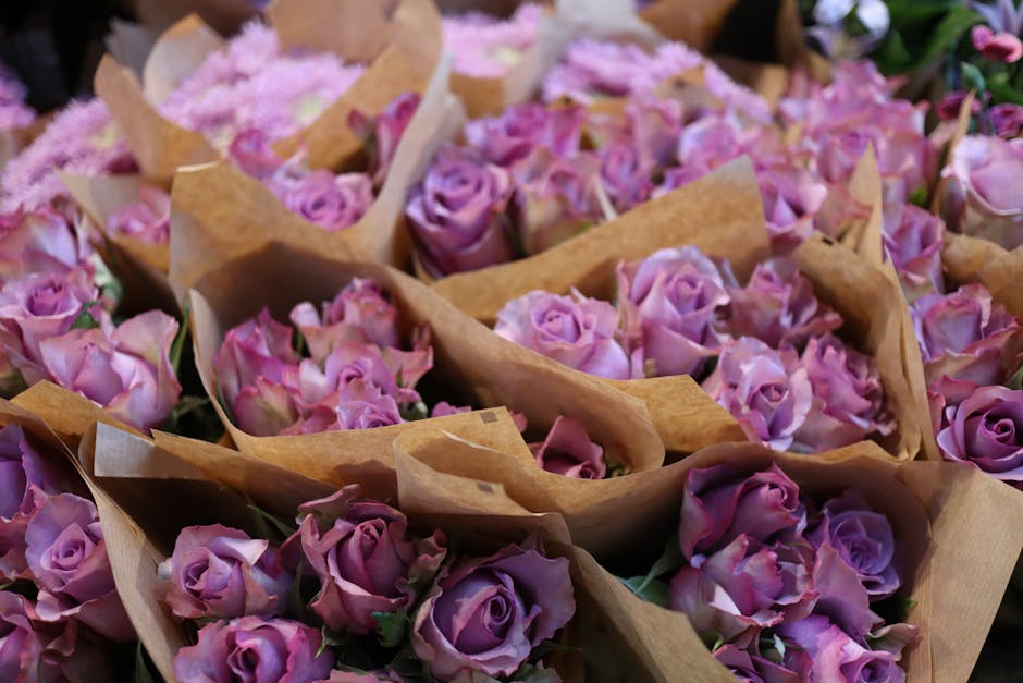 A close-up view of numerous small bouquets of fresh pink and purple roses wrapped in brown craft paper, arranged in a dense cluster. The roses exhibit velvety petals with subtle color gradients, from 