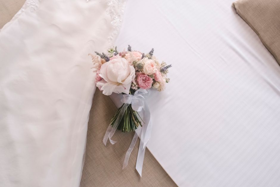 A delicate bridal bouquet featuring soft pink roses, white peonies, small lavender sprigs, and light pink lisianthus, arranged in a rounded, compact style. The flowers are fresh with vibrant petals an
